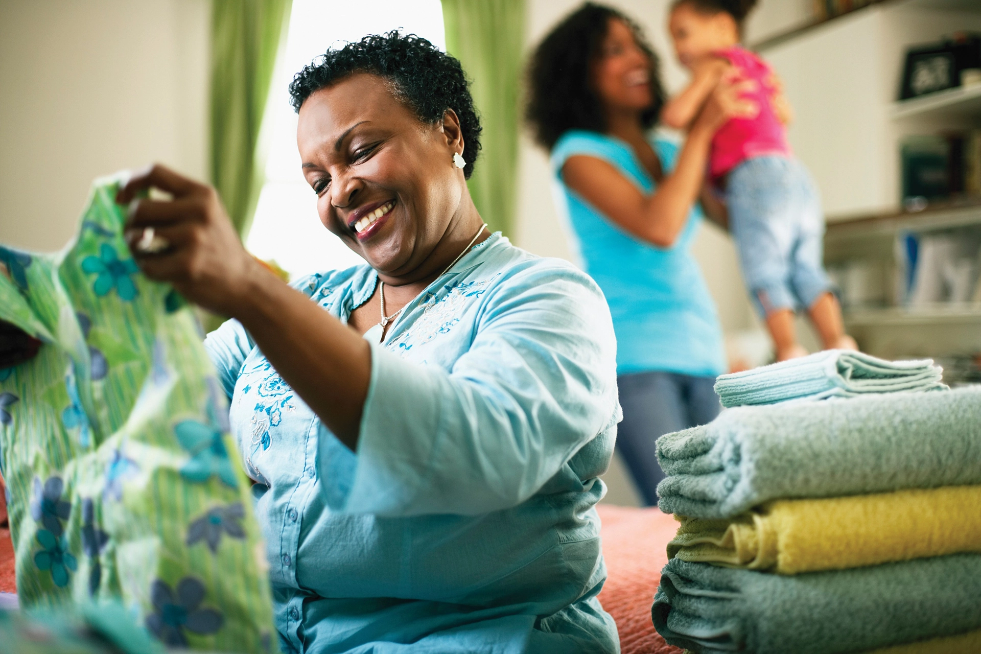 A smiling elderly woman folding laundry with a younger woman and baby in the background.